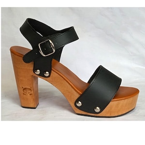 May include: A black leather sandal with a wooden platform sole and a high block heel. The shoe features a wide front strap and an adjustable ankle strap with a silver-toned metal buckle. Silver metal studs secure the leather straps to the wood base.