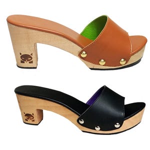 May include: Two pairs of open-toe clogs with wooden heels and soles. One pair features tan leather uppers with gold-tone studs and a bright green interior lining. The other pair has black leather uppers with silver-tone studs and a purple interior lining.