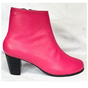 May include: A bright pink ankle boot with a smooth finish, featuring a black block heel and a black sole. The boot has a side zipper closure and white stitching along the top edge of the ankle opening, presented from a side profile view.