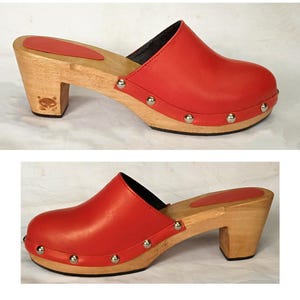 May include: A red leather clog with a wooden sole and heel. The upper is secured to the light-colored wood base with silver-toned metal studs along the perimeter. A red insole is visible on the footbed, and a small cat-shaped logo is embossed on the heel.