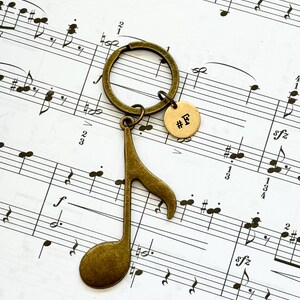 May include: A bronze-colored keychain featuring a musical eighth note pendant and a small round tag with the text "#F". The keychain is set against a backdrop of sheet music.