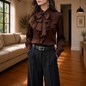 Puede incluir: Una blusa de manga larga en color marrón oscuro con un llamativo cuello de lazo con volantes en cascada y tapeta de botones frontal, combinada con pantalones negros de talle alto sujetos por un cinturón de cuero negro con hebilla plateada.