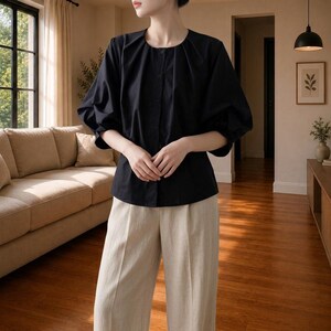 May include: A black blouse with a round neckline, a front button placket, and voluminous three-quarter length sleeves with gathered cuffs. The garment is paired with light beige trousers featuring a straight-leg cut and a pressed crease.