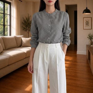 Puede incluir: Una camisa de manga larga abotonada con un patrón de cuadros vichy en blanco y negro y cuello mao, combinada con pantalones blancos de cintura alta y pernera ancha que tienen un pliegue central marcado y bolsillos laterales.