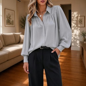 May include: A light grey long-sleeve button-down blouse with a collared neckline, gathered fabric details at the shoulders, and cuffed sleeves, paired with solid black trousers featuring a black belt with a silver buckle at the waist.
