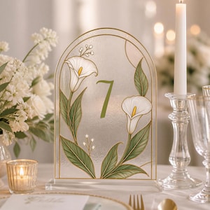 May include: An arched acrylic table number sign featuring the number 7 in a green font. The design includes white calla lily illustrations with green leaves and gold outlines, set on a clear base with a textured, frosted glass-like finish.