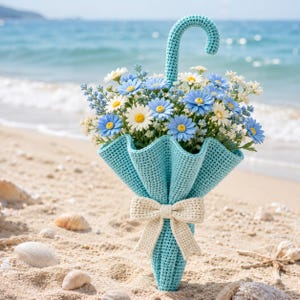 May include: A light blue crocheted umbrella-shaped container filled with a bouquet of blue and white daisy-like flowers. The umbrella features a curved handle and is tied with a cream-coloured crocheted bow around its centre section.