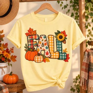 Può includere: Una t-shirt a maniche corte giallo pallido con la parola FALL in grandi lettere stampatello. Le lettere sono decorate con motivi a quadri, foglie autunnali, ghiande e un girasole. Due zucche arancioni sono posizionate alla base del testo.