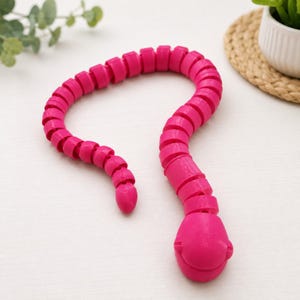 Puede incluir: Una serpiente de juguete articulada de color rosa brillante con un diseño de cuerpo segmentado. El juguete presenta una cabeza redondeada y una cola larga y flexible compuesta por múltiples anillos conectados que permiten doblar el objeto.