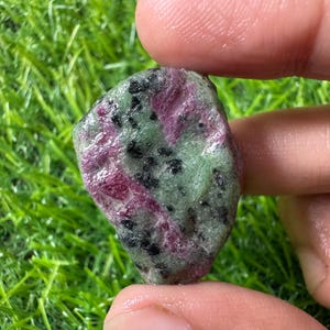 May include: A raw, unpolished ruby zoisite stone featuring a mottled texture with vibrant magenta-pink patches, light green areas, and scattered black mineral inclusions. The stone has an irregular, rounded shape with a natural, uneven surface.