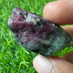 Könnte beinhalten: Ein rohes, unpoliertes Mineralexemplar mit einer Kombination aus tief magentaroten Rubinkristallen, die in eine Matrix aus undurchsichtigem, gesprenkeltem grünem Zoisit eingebettet sind, mit verstreuten schwarzen Einschlüssen im Stein.