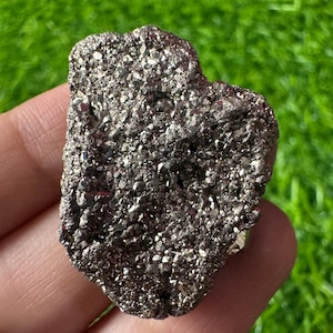 Pode incluir: Um espécime mineral de pirita metálica bruta com um aglomerado de pequenos cristais cúbicos de cor latão. A superfície da pedra é altamente reflexiva e texturizada, apresentando numerosas facetas angulares que captam a luz.