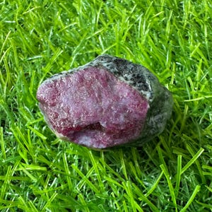 May include: A raw, unpolished mineral specimen featuring a vibrant magenta-pink crystalline centre surrounded by a dark, mottled grey and black outer crust. The stone has an irregular, jagged shape with a rough, natural texture throughout its surface.