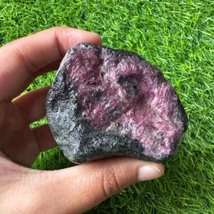 Könnte beinhalten: Ein rohes, unpoliertes Mineralexemplar mit einem tief magentafarbenen und dunkelvioletten kristallinen Zentrum, das von einer rauen, strukturierten schwarzen und dunkelgrauen Außenkruste umgeben ist. Der Stein hat eine unregelmäßige, abgerundete Form.