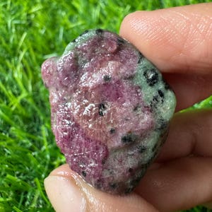 May include: A polished, irregular-shaped ruby zoisite gemstone featuring a vibrant mix of deep magenta-pink ruby crystals contrasted against a pale green zoisite matrix, speckled with small, dark black hornblende inclusions throughout the surface.