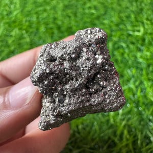 Pode incluir: Um espécime mineral de pirita metálica bruta apresentando um aglomerado de pequenos cristais cúbicos de cor latão com uma superfície reflexiva e texturizada. O mineral tem uma forma irregular e irregular com fendas profundas.