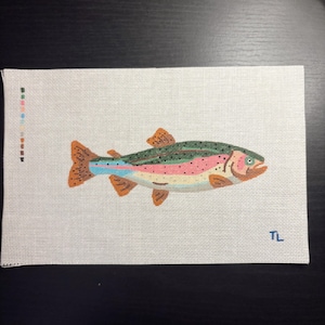May include: A rectangular needlepoint canvas featuring a colourful rainbow trout design. The fish has a green back with black spots, a pink lateral stripe, and orange fins. A vertical colour palette guide is on the left, and the initials TL are in the bottom right corner.