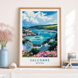 Puede incluir: Una lámina artística enmarcada que muestra un paisaje costero de Salcombe, Devon, con agua azul, veleros y casas en la ladera. El primer plano muestra flores rosas y el texto SALCOMBE DEVON está impreso en azul en la parte inferior del borde.
