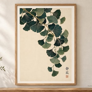 May include: A framed wall art print featuring a botanical illustration of a ginkgo branch with dark green fan-shaped leaves hanging downward against a cream background. The print includes vertical Japanese text and a small red square seal in the corner.