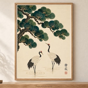 May include: A framed wall art print featuring two white cranes with black wing tips and red crowns standing in shallow water beneath a large, dark green pine tree branch. The artwork includes black Japanese calligraphy characters and a small red seal.