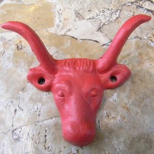 May include: Red cast iron bull head wall hook. The bull's head is facing forward with large horns curving up and out. The hook is attached to the back of the bull's head.
