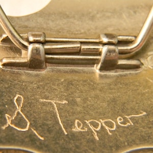 Puede incluir: Primer plano de la parte posterior de una hebilla de cintur&oacute;n de metal dorado con cierre de bisagra. La hebilla est&aacute; grabada con la firma "S. Tepper".