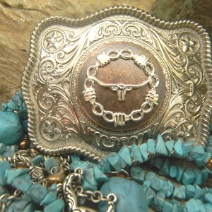 Puede incluir: Una hebilla de cintur&oacute;n de plata con un dise&ntilde;o ornamentado que presenta una cabeza de toro de color cobre rodeada de alambre de p&uacute;as. La hebilla est&aacute; sobre un lecho de piedras de color turquesa.