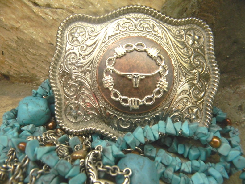 Puede incluir: Una hebilla de cintur&oacute;n de plata con un dise&ntilde;o ornamentado que presenta una cabeza de toro de color cobre rodeada de alambre de p&uacute;as. La hebilla est&aacute; sobre un lecho de piedras de color turquesa.