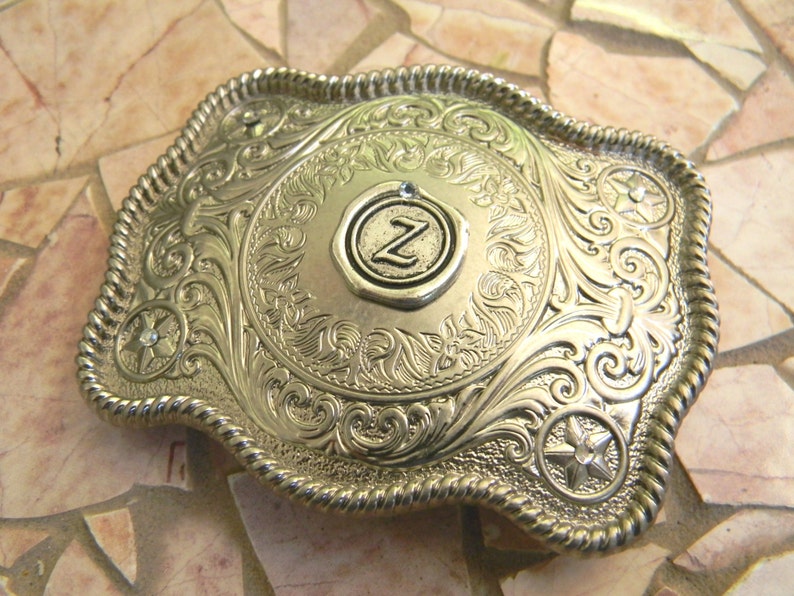 Puede incluir: Hebilla de cintur&oacute;n de plata con un dise&ntilde;o ornamentado y una letra "Z" en relieve en el centro.