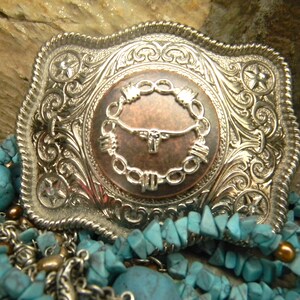 Puede incluir: Hebilla de cintur&oacute;n de plata con un dise&ntilde;o ornamentado que presenta una cabeza de toro de color cobre dentro de un c&iacute;rculo de alambre de p&uacute;as. La hebilla est&aacute; rodeada de piedras turquesa y cadenas de plata.