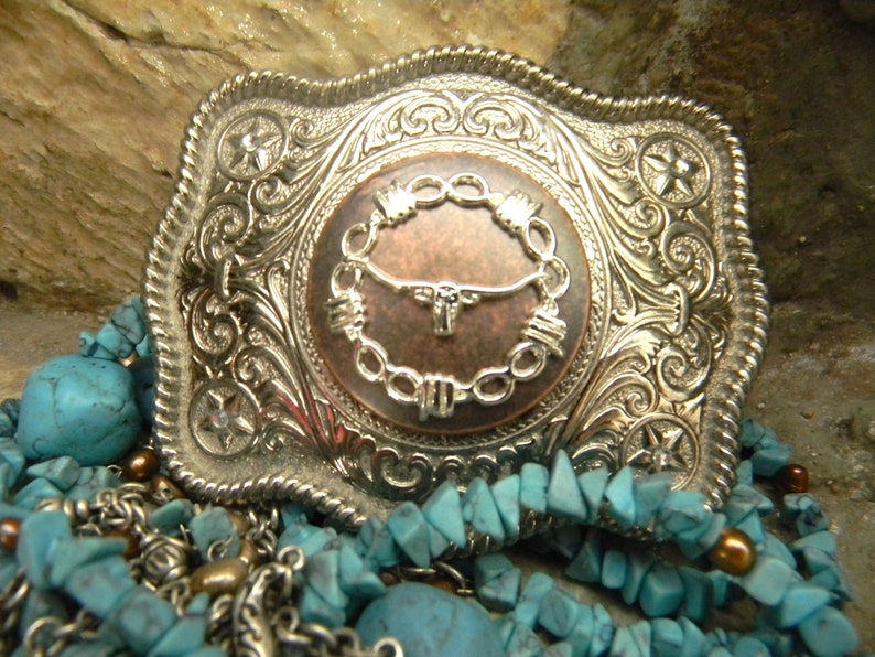 Puede incluir: Hebilla de cintur&oacute;n de plata con un dise&ntilde;o ornamentado que presenta una cabeza de toro de color cobre dentro de un c&iacute;rculo de alambre de p&uacute;as. La hebilla est&aacute; rodeada de piedras turquesa y cadenas de plata.