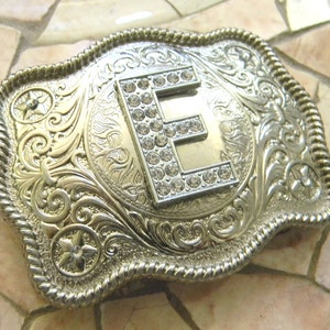 May include: Silver belt buckle with an ornate design and a large letter E in the center, decorated with rhinestones.