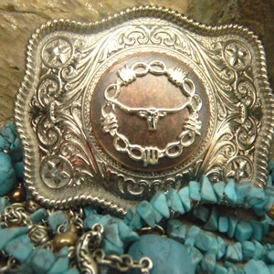 Puede incluir: Una hebilla de cintur&oacute;n de plata con un dise&ntilde;o ornamentado que presenta un centro de color cobre con un c&iacute;rculo de alambre de p&uacute;as y una cabeza de toro. La hebilla est&aacute; rodeada de piedras turquesa y cadenas de plata.