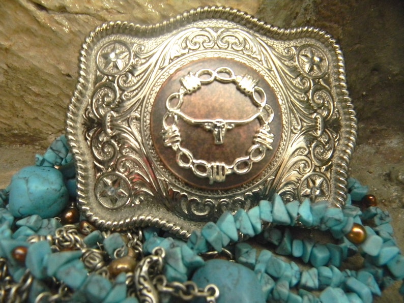 Puede incluir: Una hebilla de cintur&oacute;n de plata con un dise&ntilde;o ornamentado que presenta un centro de color cobre con un c&iacute;rculo de alambre de p&uacute;as y una cabeza de toro. La hebilla est&aacute; rodeada de piedras turquesa y cadenas de plata.