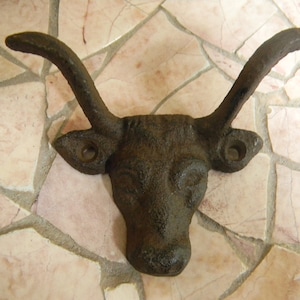 Peut inclure: Un crochet mural en forme de tête de taureau en fonte avec deux cornes. Le crochet est brun foncé et a une finition rustique.