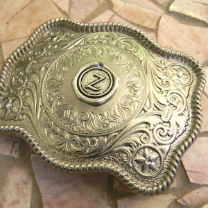Puede incluir: Hebilla de cintur&oacute;n de plata con un dise&ntilde;o ornamentado y una letra "Z" en relieve en el centro.
