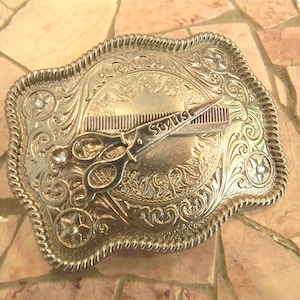 May include: Silver-coloured metal belt buckle with an ornate floral design. The buckle features a comb and scissors, with the word "Stylist" on the scissors. The edges are rope-like, and the buckle has star-shaped accents with clear rhinestones.