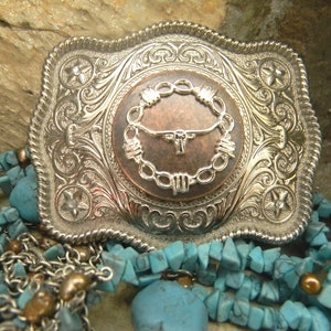 Puede incluir: Una hebilla de cintur&oacute;n de plata con un dise&ntilde;o ornamentado que presenta una cabeza de toro de color cobre dentro de un c&iacute;rculo de alambre de p&uacute;as. La hebilla est&aacute; rodeada de piedras turquesa.