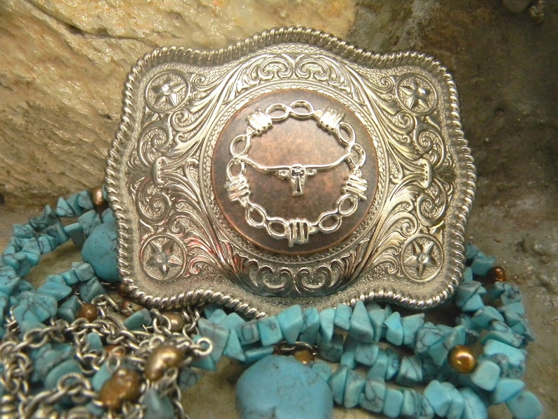 Puede incluir: Una hebilla de cintur&oacute;n de plata con un dise&ntilde;o ornamentado que presenta una cabeza de toro de color cobre dentro de un c&iacute;rculo de alambre de p&uacute;as. La hebilla est&aacute; rodeada de piedras turquesa.