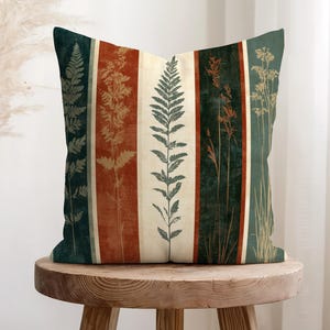 May include: A square throw pillow featuring a vertical striped pattern with alternating bands of dark green, burnt orange, and cream. The design displays botanical silhouettes of ferns and tall grasses printed in contrasting colours across the stripes.