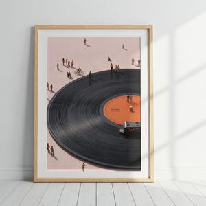 May include: A framed art print featuring a large black vinyl record on a light pink background. Tiny figures of people are walking and standing on the surface of the record and the surrounding area. A small record player arm rests on the orange center label.