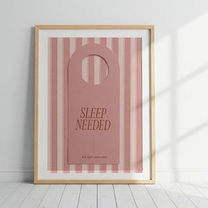 Può includere: Una stampa incorniciata con la forma di un cartello da porta rosa polvere centrato su uno sfondo a righe verticali rosa polvere e bianco. Il cartello riporta il testo SLEEP NEEDED in un carattere serif scuro e DO NOT DISTURB in basso.