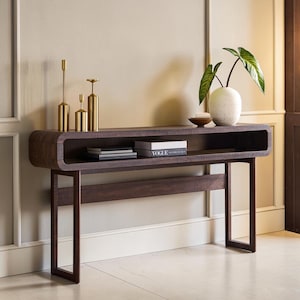 May include: A dark wood console table with a rectangular open shelf, featuring three gold-toned metal candle holders, a white speckled ceramic vase with a green plant, and two stacked books with one labeled Vogue on the top surface.