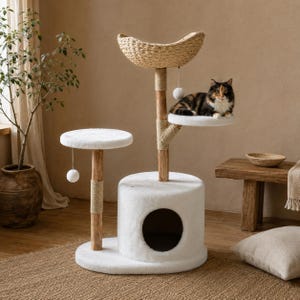 May include: A multi-level cat tree featuring a white plush-covered base with a circular entrance, two white plush platforms, and a woven seagrass basket at the top. A calico cat rests on the upper platform, and two white pom-pom toys hang down.