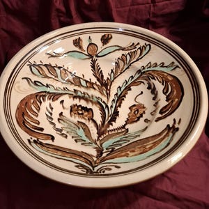 May include: A round ceramic bowl with a cream-colored base featuring hand-painted brown and light blue floral motifs. The design includes symmetrical, sweeping leaf-like patterns radiating from the center, framed by thin brown circular lines.