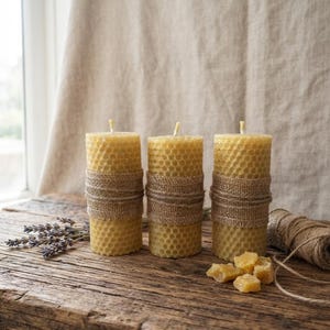 May include: Three cylindrical yellow beeswax candles with a textured honeycomb pattern, each wrapped with a strip of tan burlap fabric and twine. Small pieces of raw yellow wax and dried lavender sprigs are placed near the candles.