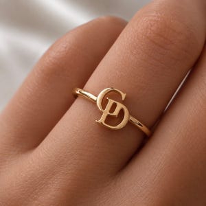 May include: A gold-toned ring featuring a polished metal band and a central decorative element composed of the interlocking capital letters G and P. The design is smooth and reflective, showcasing a continuous metallic finish throughout the piece.