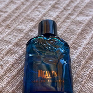Peut inclure: Un petit flacon en verre bleu translucide de Chopard Heaven Eau de Toilette. Le flacon présente un bouchon cylindrique noir et un motif de feuille moulé sur la partie supérieure avant. Le texte HEAVEN, Chopard et EAU DE TOILETTE est doré.