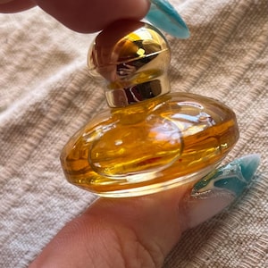 Peut inclure: Un petit flacon de parfum en verre ambré, de forme ronde, doté d'un bouchon sphérique lisse et réfléchissant de couleur dorée. Le flacon présente une base large et aplatie ainsi qu'un col étroit, contenant un liquide doré translucide.