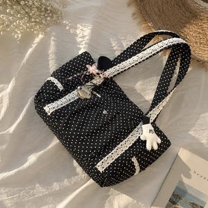 Puede incluir: Un bolso tote negro con estampado de lunares blancos, con ribete de encaje blanco a lo largo de las asas y el panel frontal. El bolso incluye un peque&ntilde;o dije plateado, un detalle de cinta rosa y un llavero con forma de guante blanco.